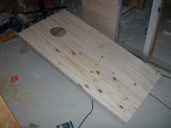Unfinished DIY cornhole board with hole cut and folding legs on a garage floor